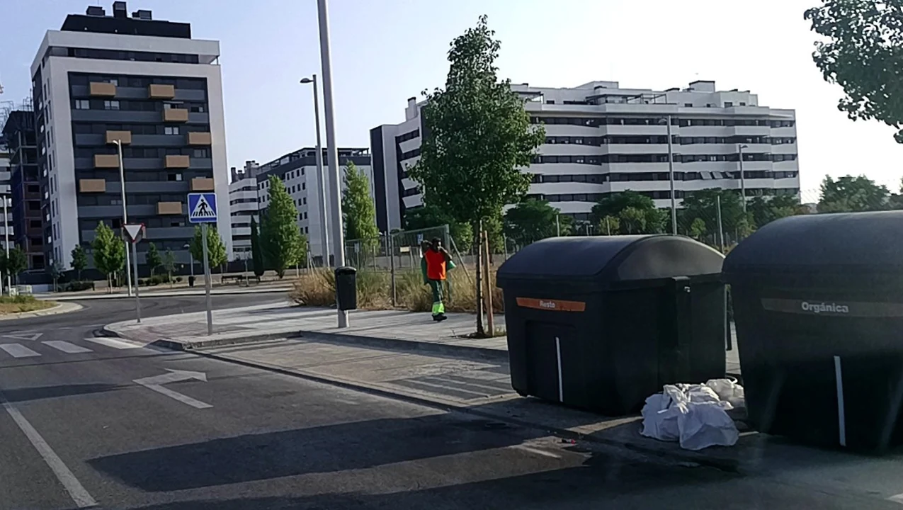 Escena urbana con contenedores, bolsas en el suelo y un operario al fondo